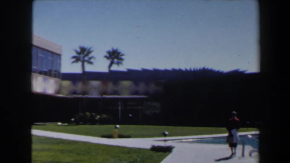 Las Vegas 1958 exterior with palm trees, lawn, shaded building, pool edge, and a small distant figure near the water.