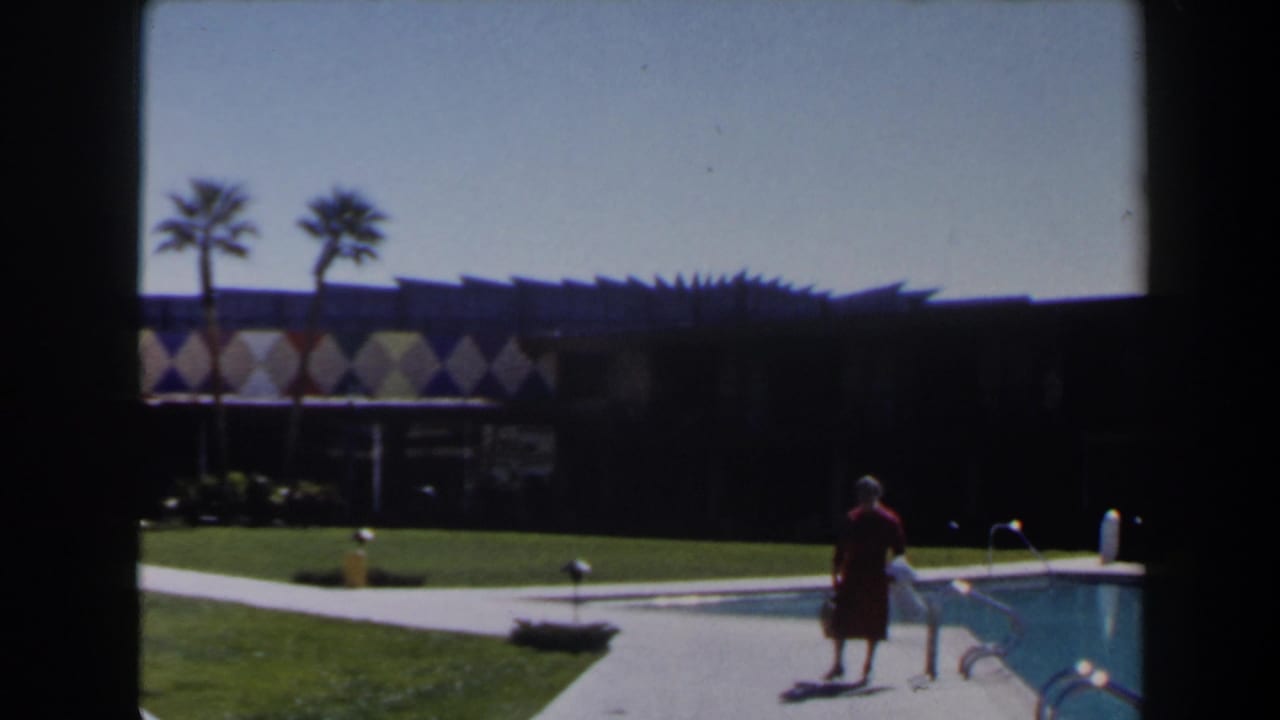 Woman in a red dress walks beside a swimming pool at a sunny resort.