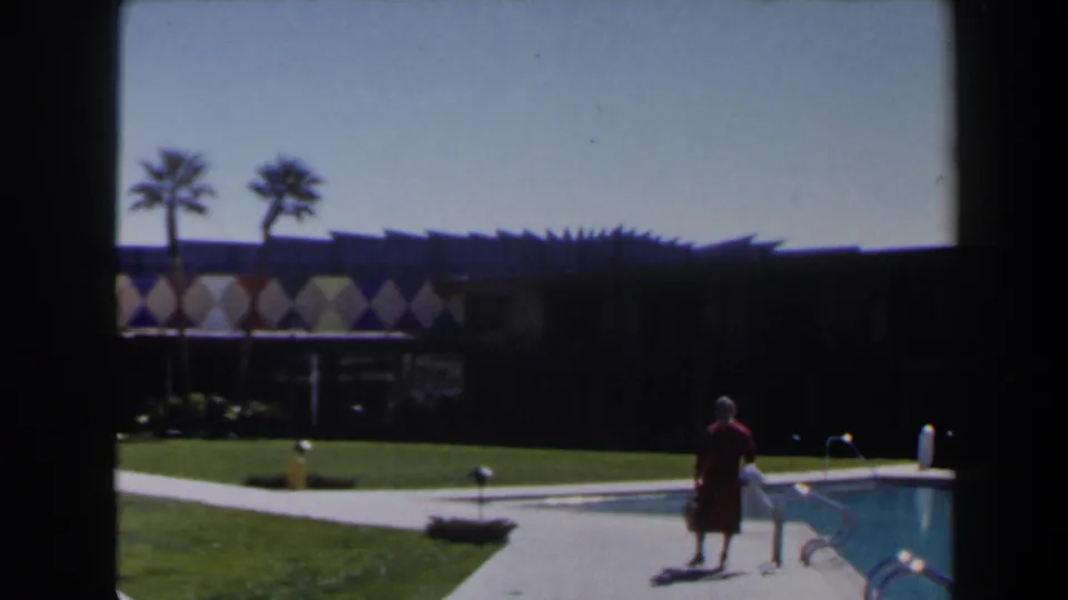 In 1958 Las Vegas home-movie footage, a woman in a red dress walks along a pool deck near lawn, palms, and a geometric resort facade.