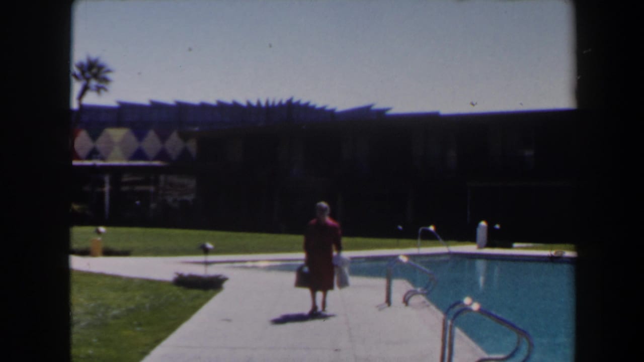 A woman in a red dress carries bags near a turquoise pool, with palm trees, lawn, and a dark mid-century resort building behind her.
