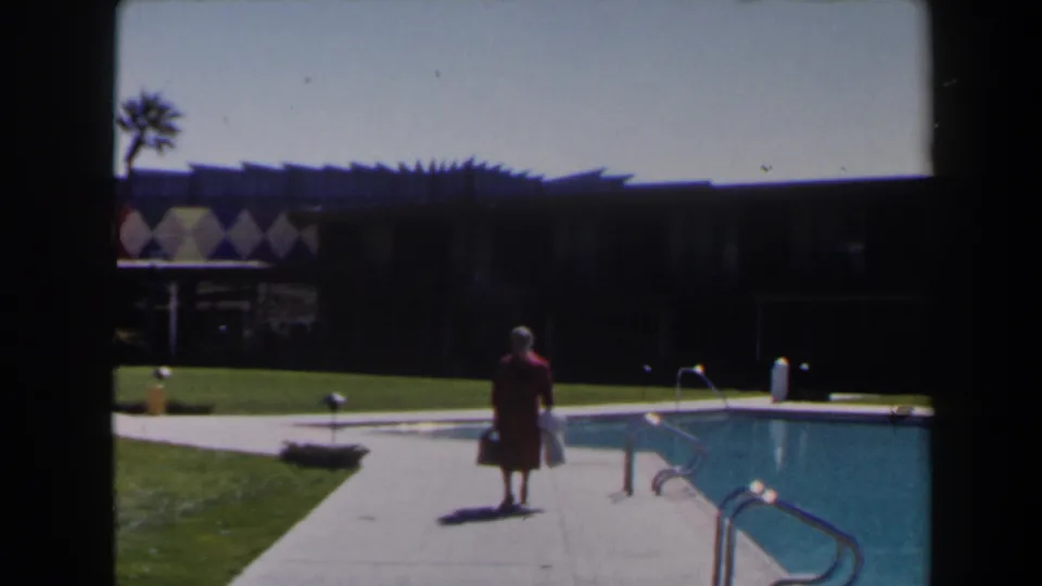 A woman in a red dress carries bags near a turquoise pool, with palm trees, lawn, and a dark mid-century resort building behind her.