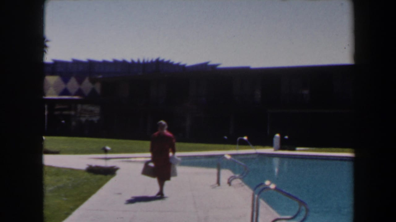 The woman in red moves closer along the pool deck in Las Vegas, with the bright pool, metal ladder, lawn, and shaded resort facade visible.