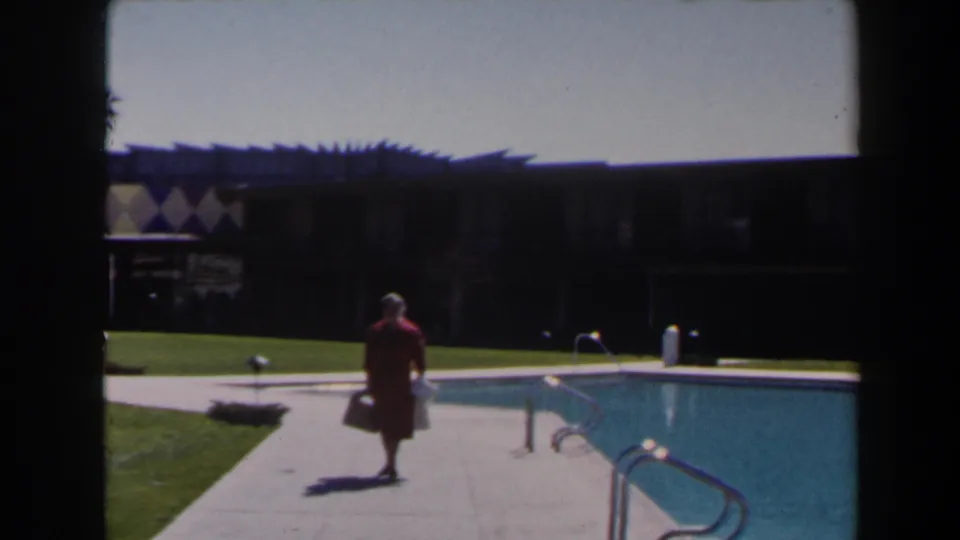 The woman in red moves closer along the pool deck in Las Vegas, with the bright pool, metal ladder, lawn, and shaded resort facade visible.