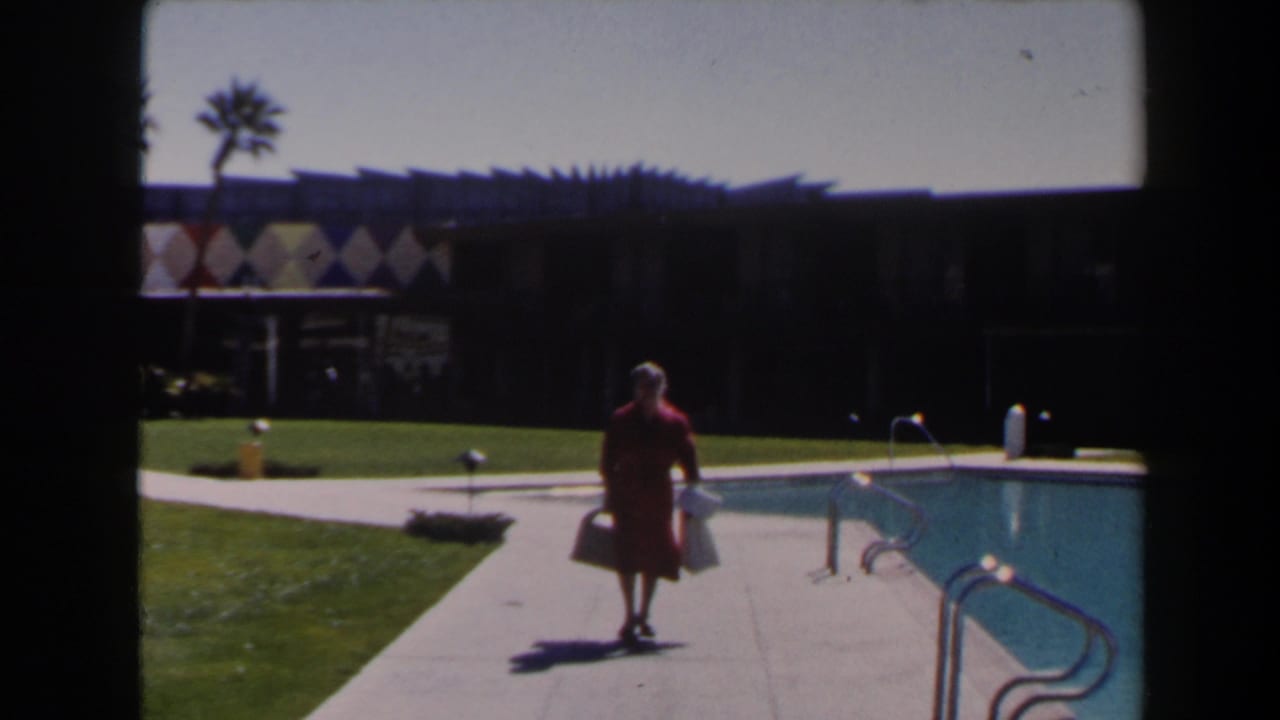 Near the end of the clip, the woman in red passes the pool area as the diamond-patterned resort facade, palms, lawn, and railings remain in view.