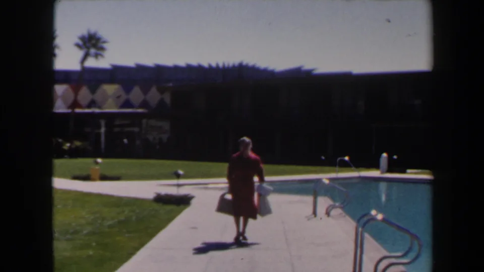 Near the end of the clip, the woman in red passes the pool area as the diamond-patterned resort facade, palms, lawn, and railings remain in view.