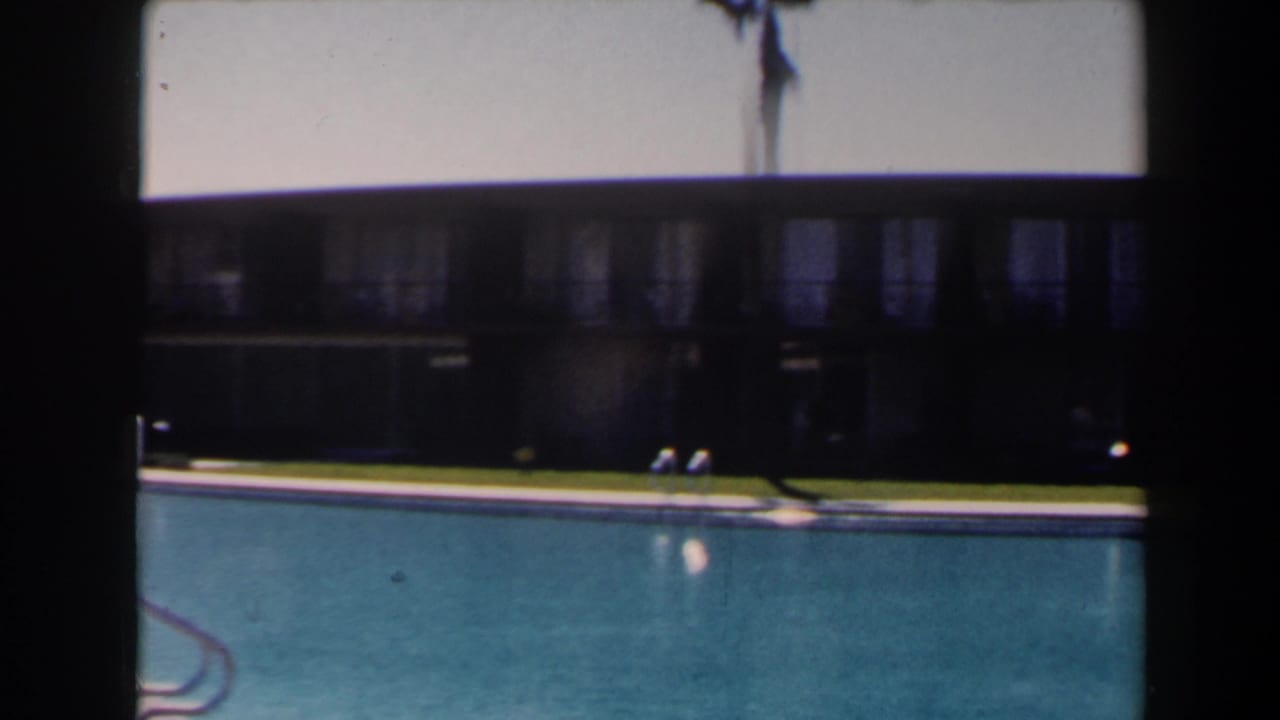 1958 Las Vegas hotel pool area with blue water, a metal ladder, lawn, and dark motel balcony building beyond.