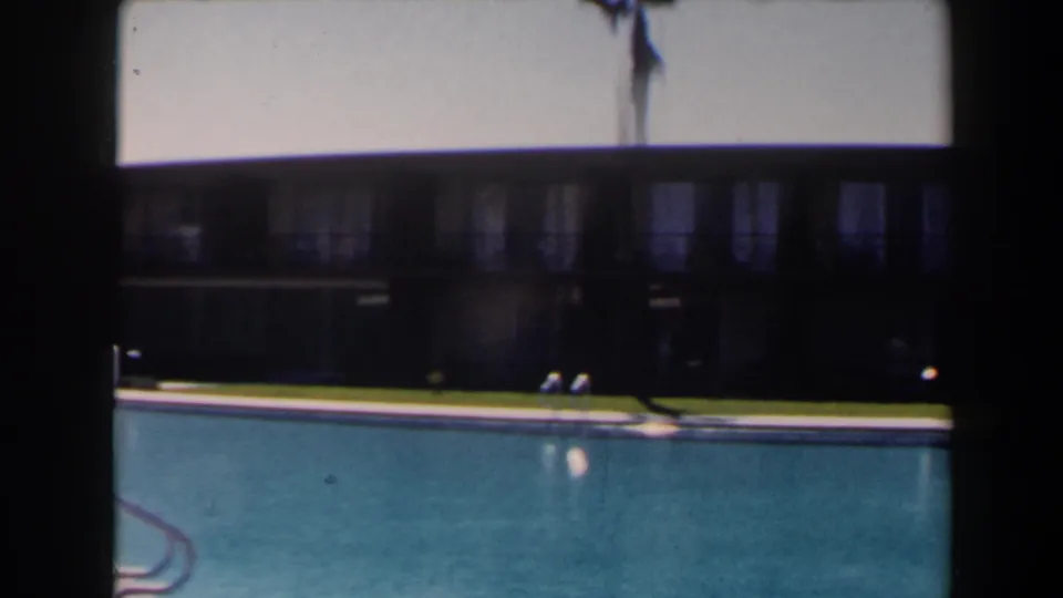 1958 Las Vegas hotel pool area with blue water, a metal ladder, lawn, and dark motel balcony building beyond.