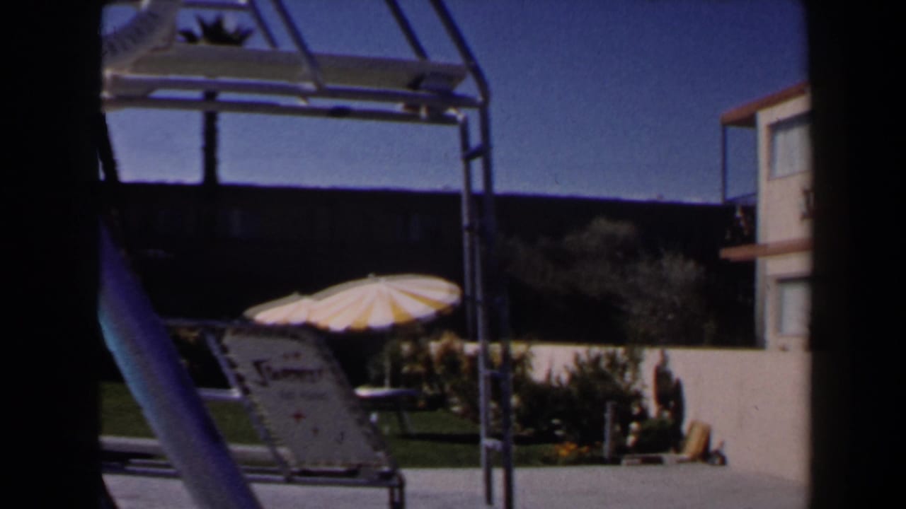 1958 Las Vegas pool courtyard view with a metal pool structure, reclining lounge chair, striped umbrella, lawn, and motel facade.