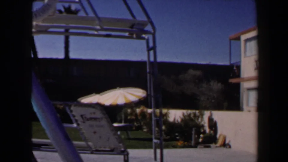 1958 Las Vegas pool courtyard view with a metal pool structure, reclining lounge chair, striped umbrella, lawn, and motel facade.