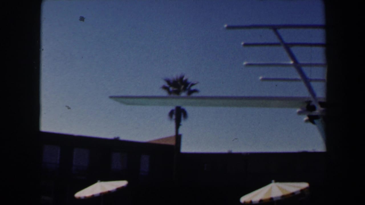 1958 Las Vegas poolside frame with the diving board crossing the sky above a palm tree, striped parasols, and resort building.