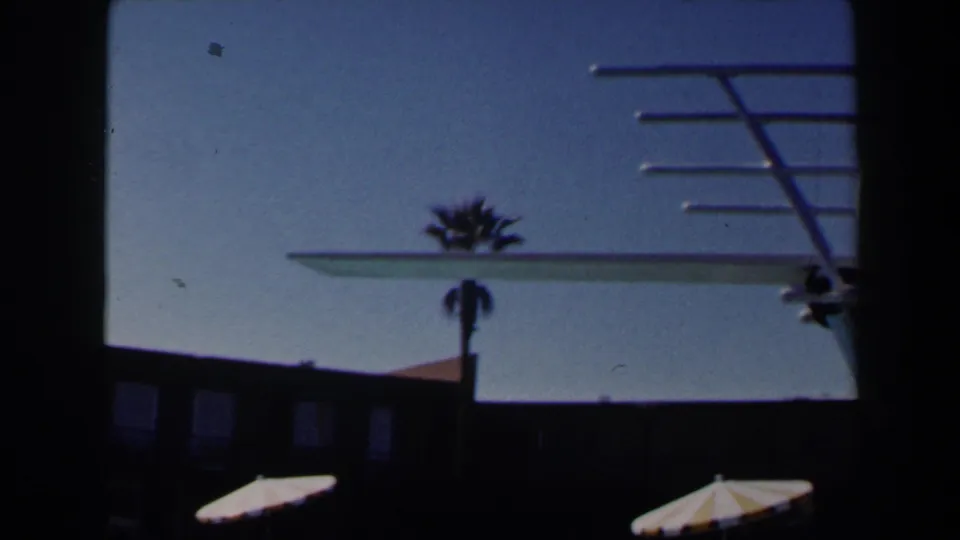 1958 Las Vegas poolside frame with the diving board crossing the sky above a palm tree, striped parasols, and resort building.