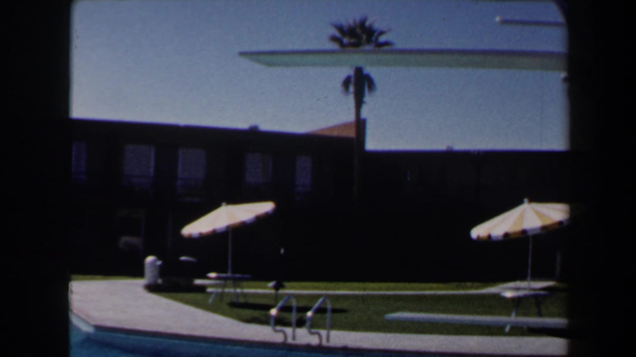 Las Vegas resort pool scene with striped parasols on the lawn, a pool edge, diving board, palm tree, and shadowed building.