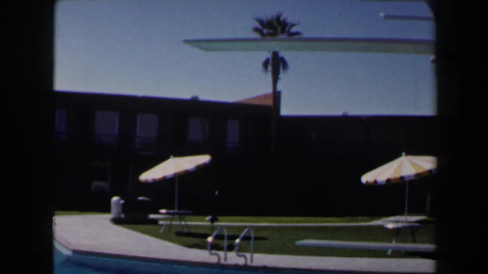 Las Vegas resort pool scene with striped parasols on the lawn, a pool edge, diving board, palm tree, and shadowed building.