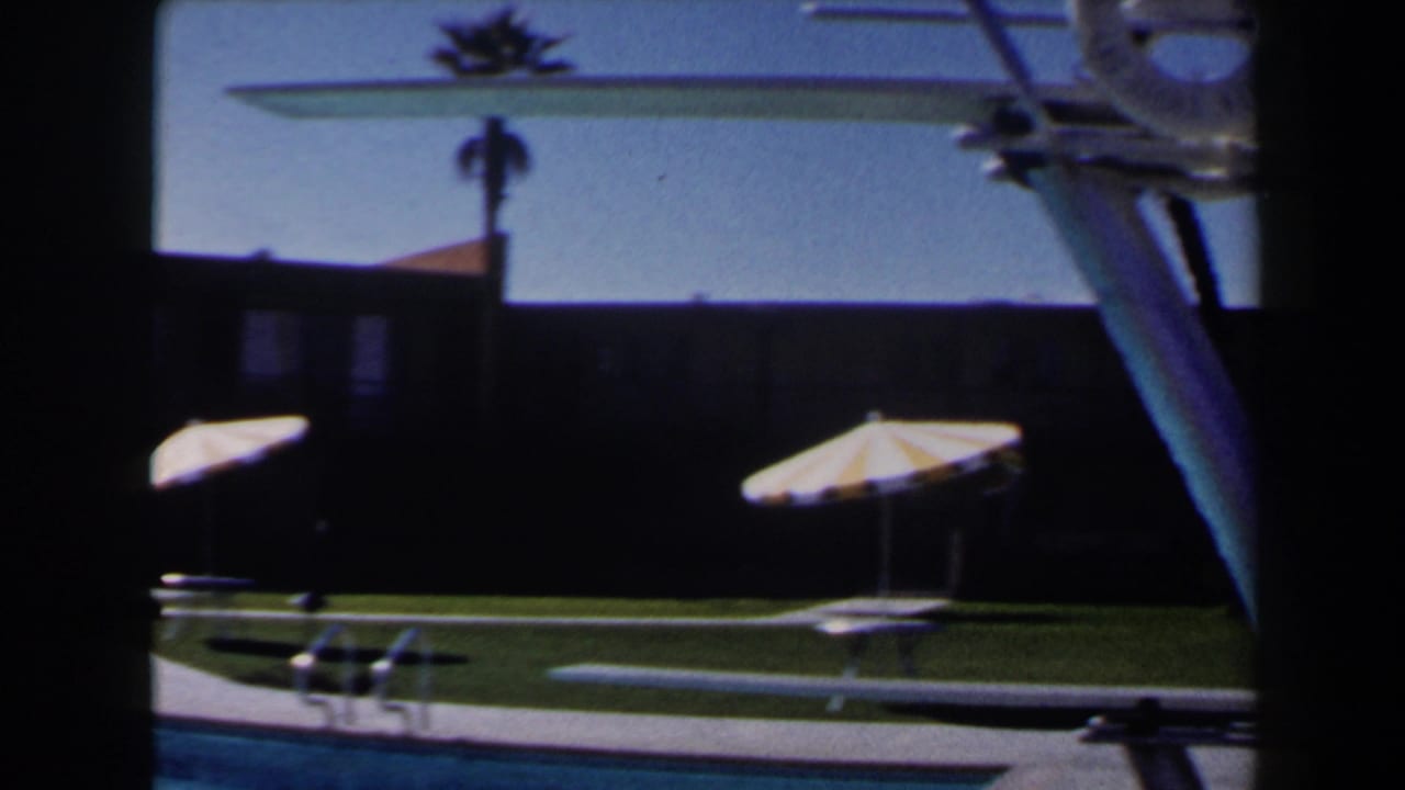 Poolside scene with striped umbrellas, diving board, lawn, and dark building facade.