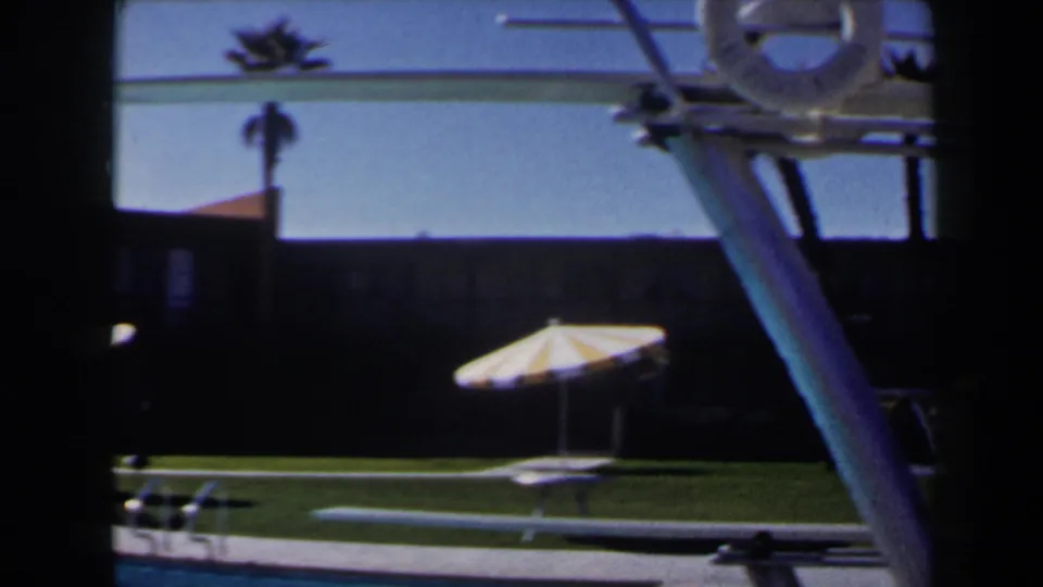 1958 Las Vegas poolside frame with a striped umbrella, diving board structure, palm silhouettes, and dark motel-like facade.