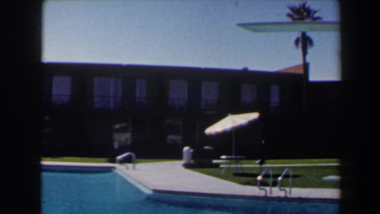 1958 Las Vegas 8mm frame showing a pool corner, ladder, striped umbrella, lounge furniture, lawn, and shaded building facade.