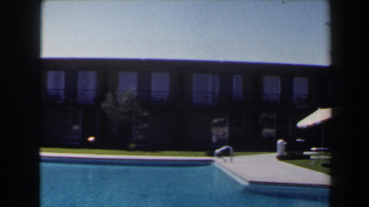 1958 Las Vegas poolside frame showing blue pool water, a deck corner, ladder, striped umbrella, lawn, and motel-like facade.