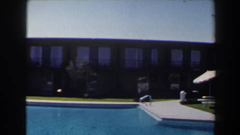 1958 Las Vegas poolside frame showing blue pool water, a deck corner, ladder, striped umbrella, lawn, and motel-like facade.