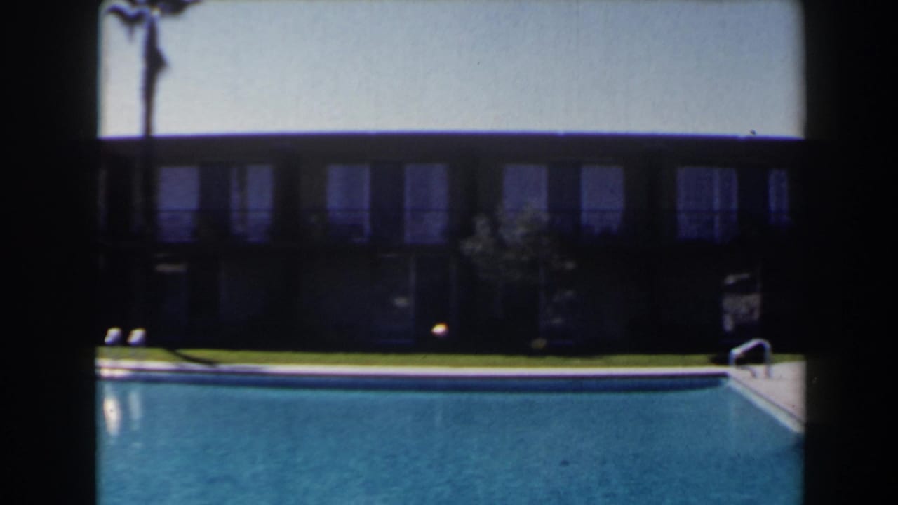 1958 Las Vegas home-movie frame showing a blue outdoor pool, lawn, palm trunk, and long windowed lodging building.