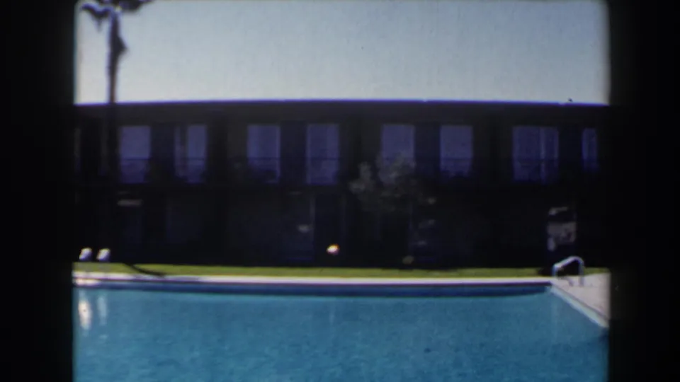 1958 Las Vegas home-movie frame showing a blue outdoor pool, lawn, palm trunk, and long windowed lodging building.