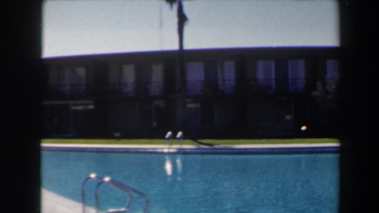 1958 Las Vegas frame of a motel or apartment pool area with metal ladder, blue water, lawn, and shaded building facade.