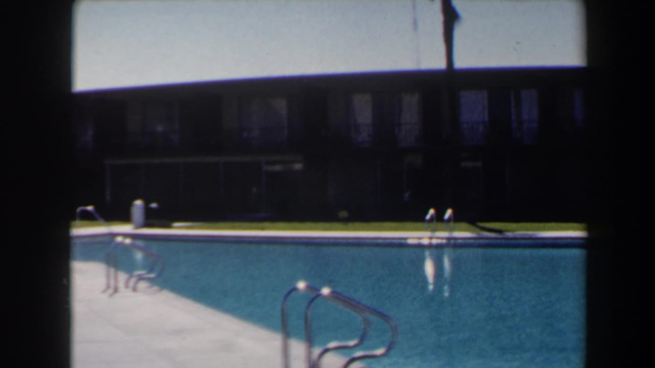 1958 Las Vegas home-movie still showing pool deck, blue water, multiple ladders, lawn, and a low windowed building.