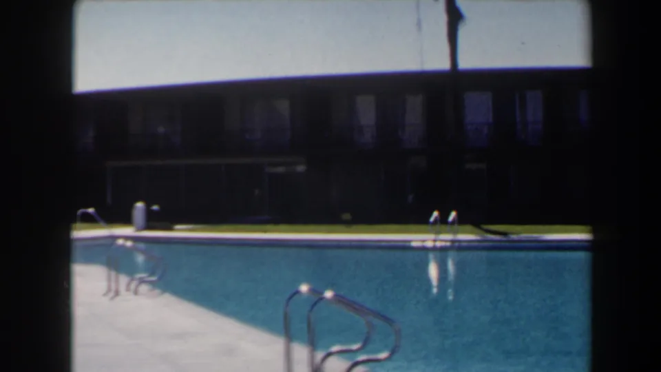 1958 Las Vegas home-movie still showing pool deck, blue water, multiple ladders, lawn, and a low windowed building.