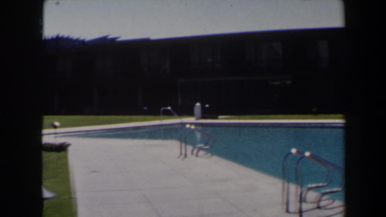 1958 Las Vegas frame showing a wide motel or apartment pool scene with concrete deck, blue water, metal ladders, and shaded facade.
