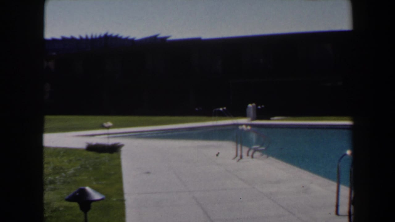 1958 Las Vegas home-movie frame showing a blue motel pool, concrete deck, green lawn, and pool ladder in bright sun.
