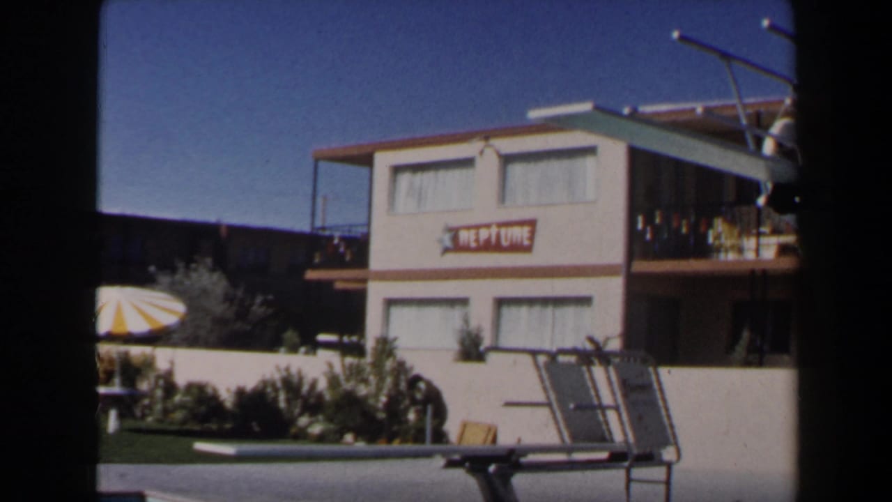 1958 Las Vegas frame of a motel pool area with a NEPTUNE sign, striped umbrella, diving board, and two-story lodging building.
