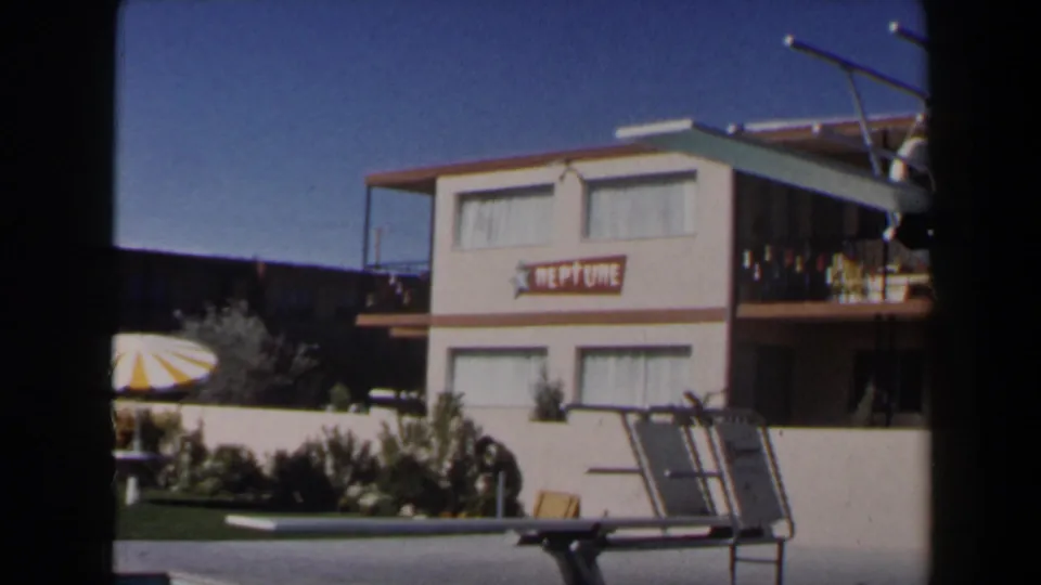 1958 Las Vegas frame of a motel pool area with a NEPTUNE sign, striped umbrella, diving board, and two-story lodging building.