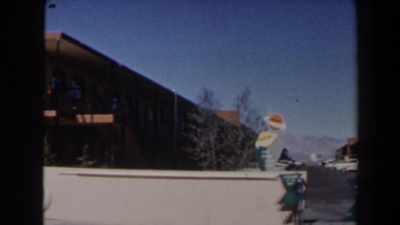 1958 Las Vegas frame with a low motel building, novelty hamburger-and-drink sign, driveway, parked cars, and mountains beyond.
