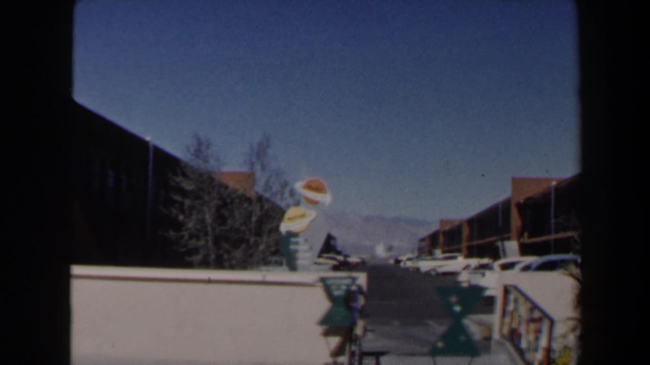 1958 Las Vegas motel view with the novelty food sign, white wall, parked cars, low buildings, and distant mountains under a clear sky.