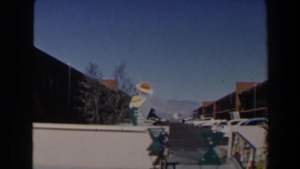 1958 Las Vegas motel view with the novelty food sign, white wall, parked cars, low buildings, and distant mountains under a clear sky.