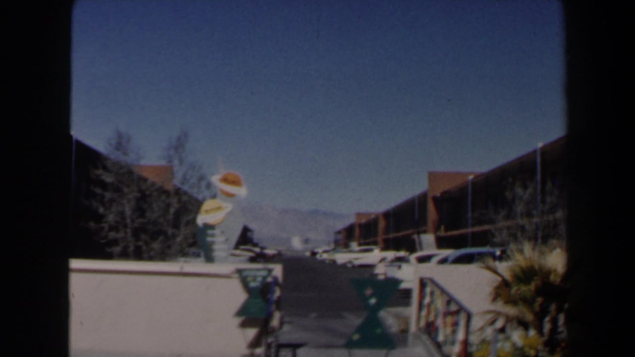 1958 Las Vegas home-movie frame looking along a motel drive toward parked cars, novelty signage, desert planting, and distant mountains.