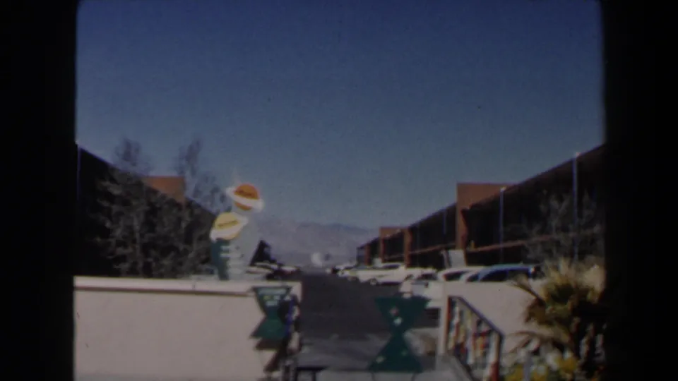 1958 Las Vegas home-movie frame looking along a motel drive toward parked cars, novelty signage, desert planting, and distant mountains.
