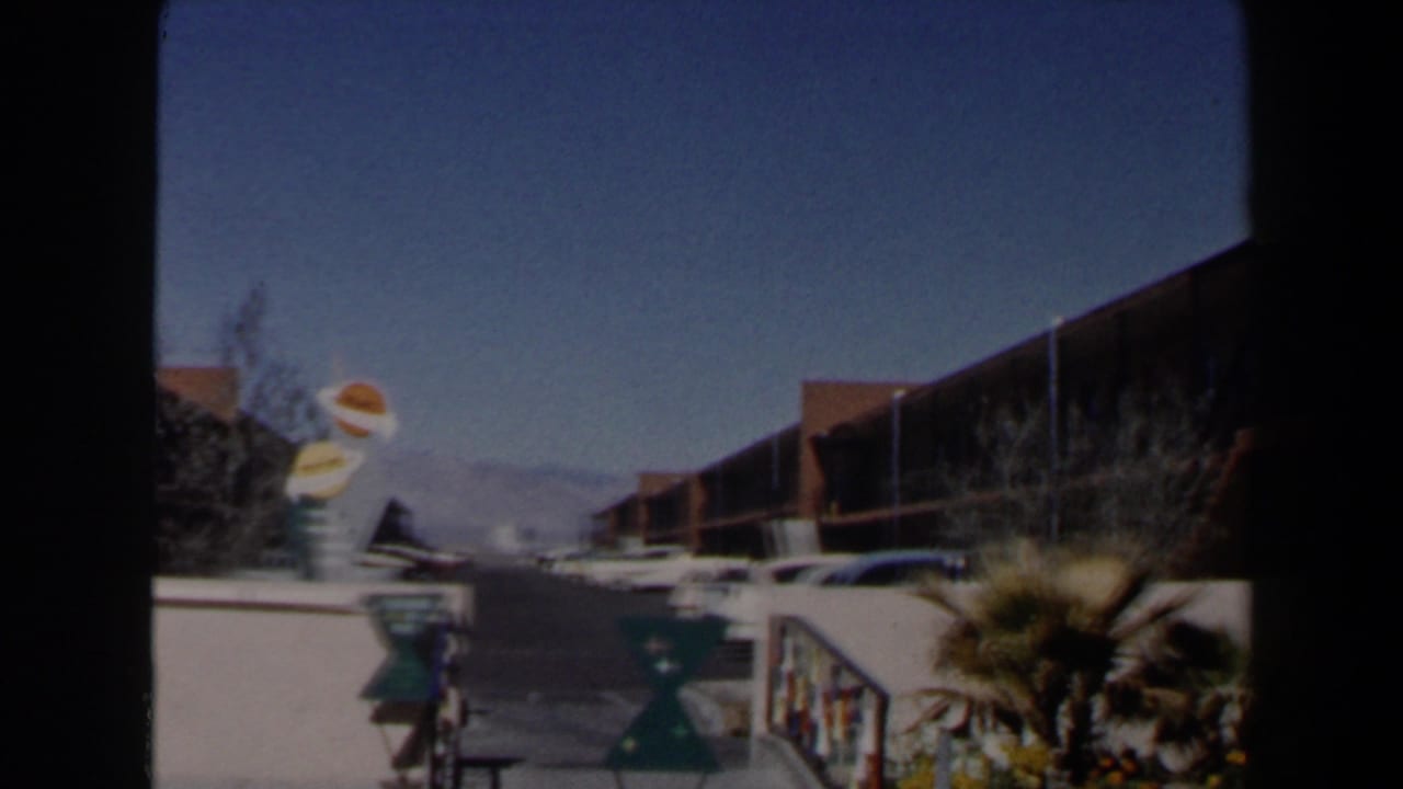 1958 Las Vegas 8mm frame showing a sunny two-story motel exterior with balconies, cars, palm plantings, and desert mountains.
