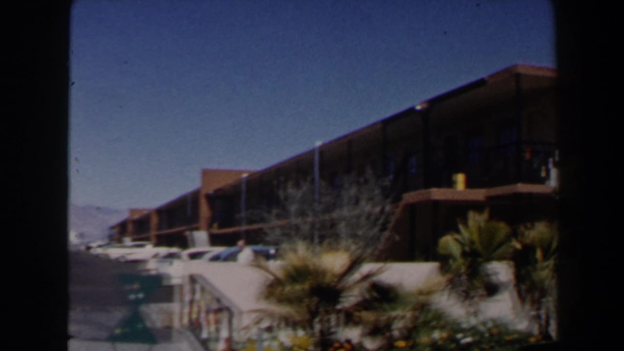 1958 Las Vegas motel exterior seen from the parking area, with balcony railings, cars, palm landscaping, and clear sky.