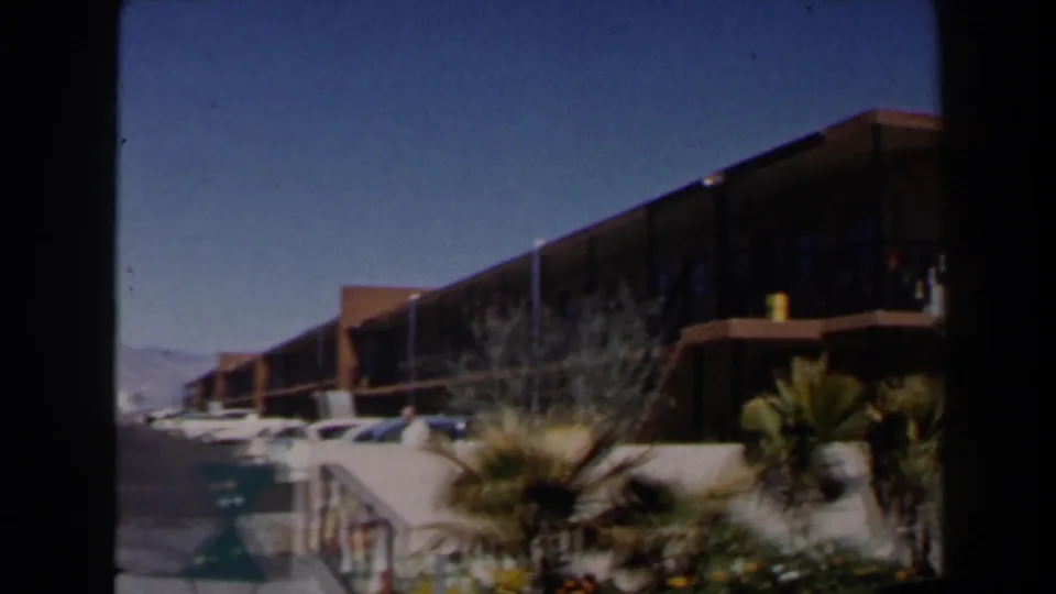 1958 Las Vegas motel exterior seen from the parking area, with balcony railings, cars, palm landscaping, and clear sky.