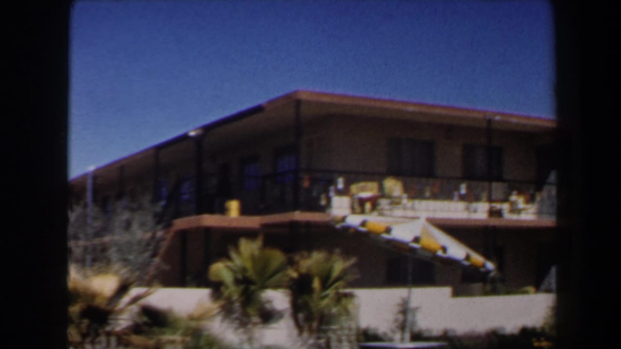 Closer 1958 Las Vegas motel view showing upper balcony rooms, striped awnings, palm plants, and a sunlit exterior wall.