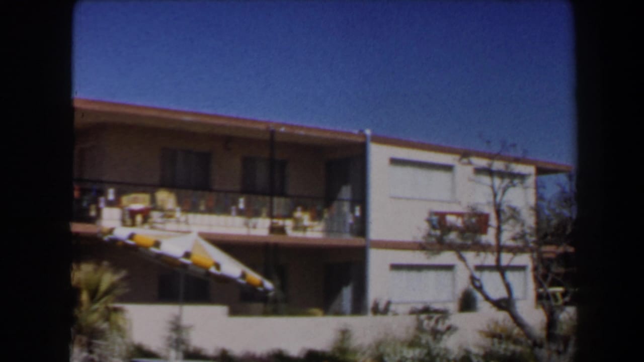 1958 Las Vegas motel corner with balcony railings, striped awnings, palm landscaping, a bare tree, and blue sky.