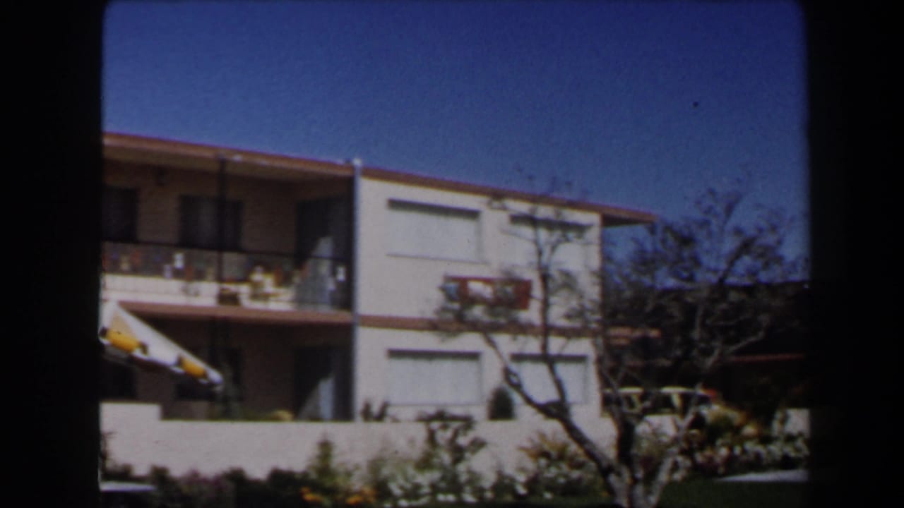 1958 Las Vegas home-movie frame showing a two-story stucco residential building with balcony, red-tiled eaves, shrubs, and clear sky.