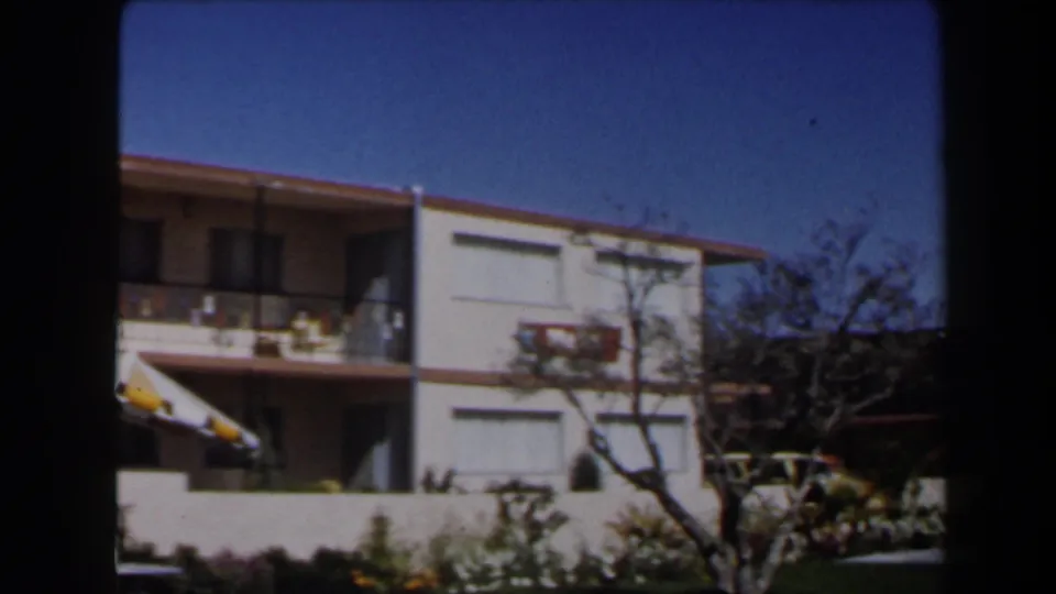 1958 Las Vegas home-movie frame showing a two-story stucco residential building with balcony, red-tiled eaves, shrubs, and clear sky.