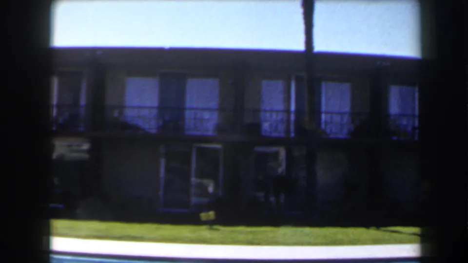 1958 Las Vegas home-movie frame of a shaded motel-style exterior beyond a lawn and bright pool edge.