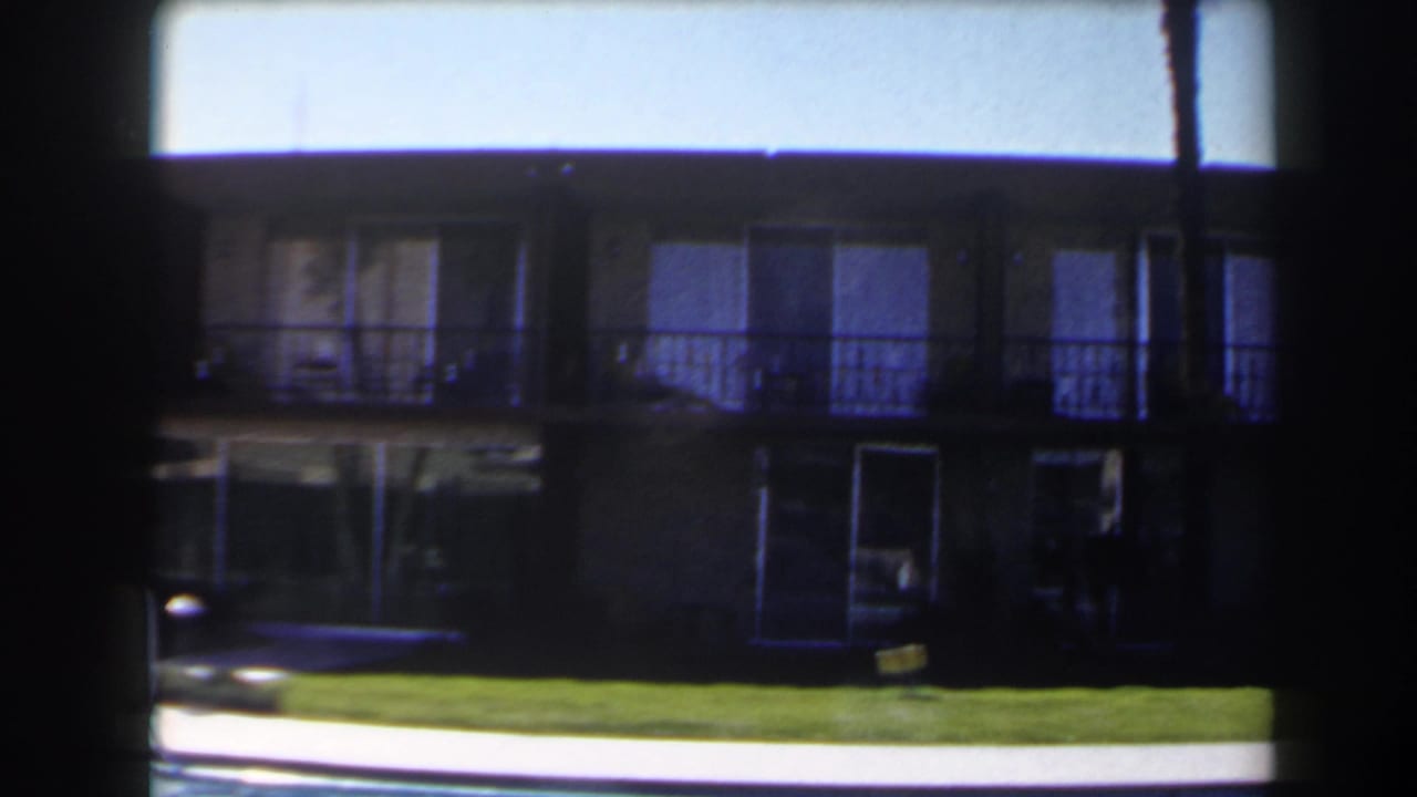 Two-story building seen across a lawn and pool edge in bright sun.