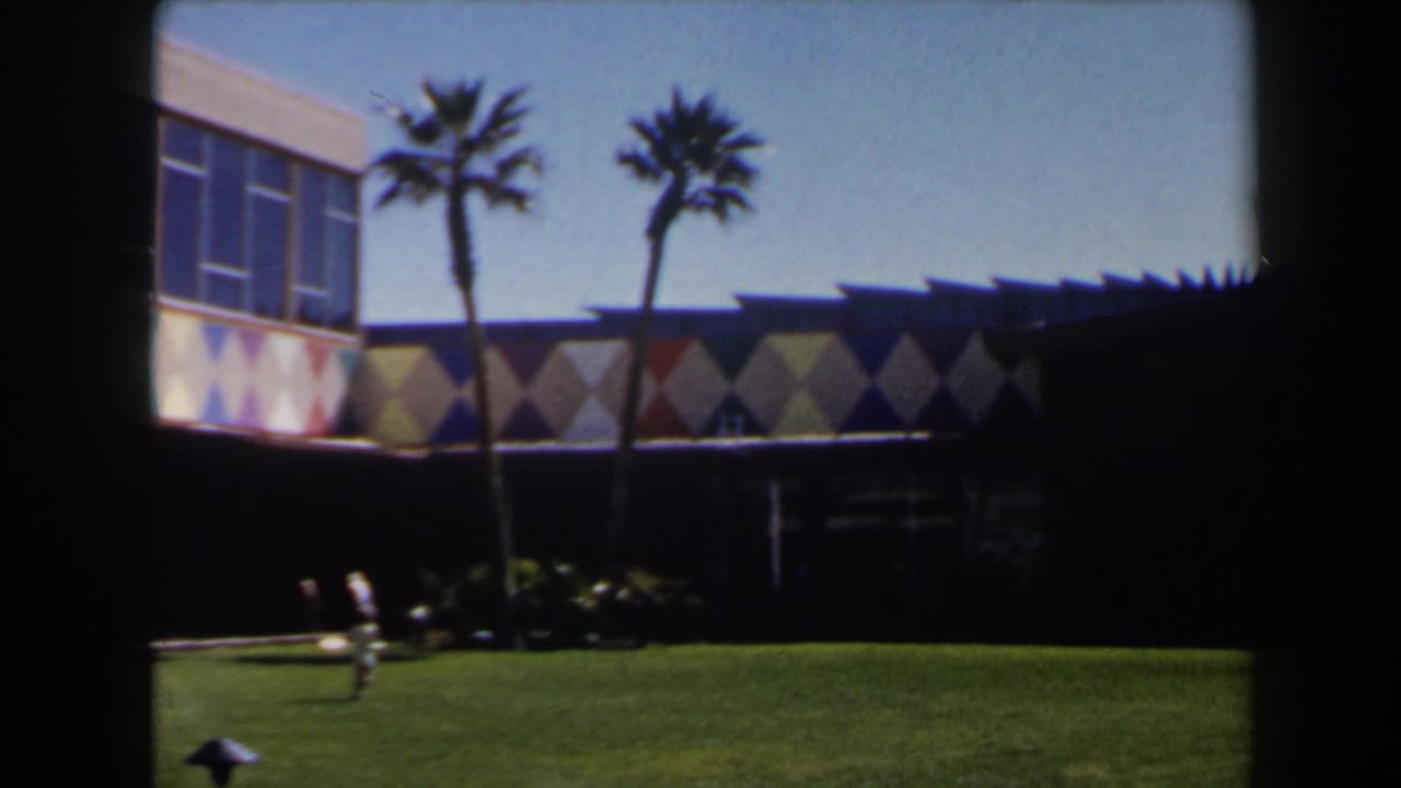 1958 Las Vegas exterior with pedestrians crossing a green lawn below palm trees and a colorful mid-century facade.