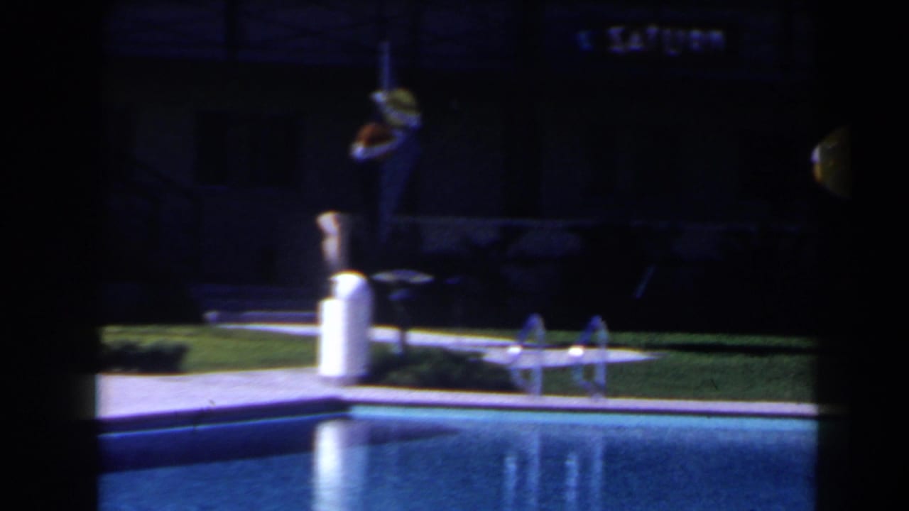 Dark-framed view across a hotel swimming pool toward lawn, chairs and a standing adult near the pool edge.
