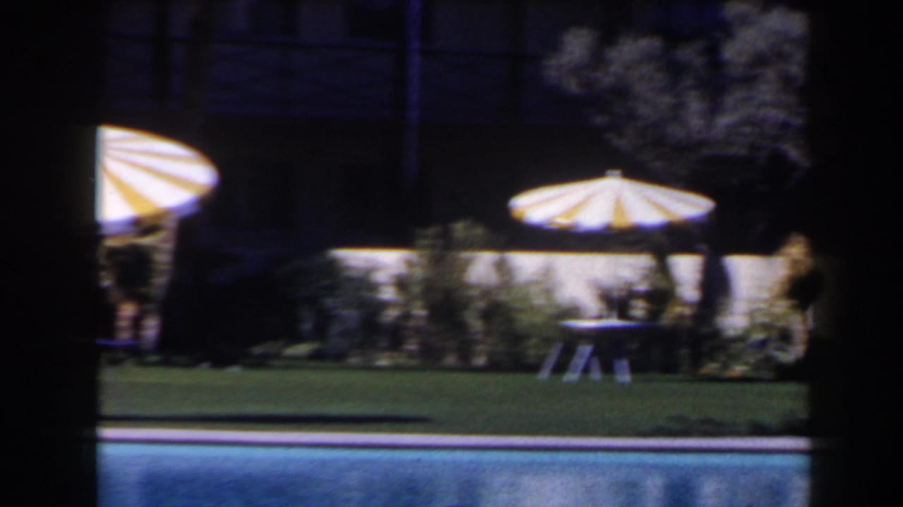 1958 Las Vegas resort poolside view with yellow-and-white striped parasols, lawn, tables and shrubs beyond the pool.