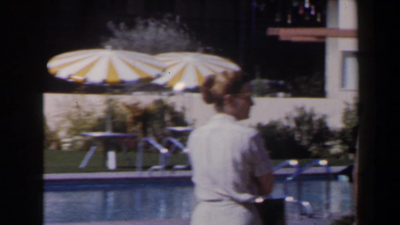 1958 Las Vegas poolside frame with two adult guests passing in front of a striped parasol, patio furniture and hotel facade.