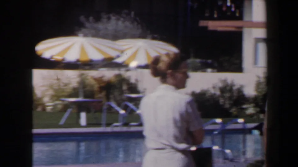1958 Las Vegas poolside frame with two adult guests passing in front of a striped parasol, patio furniture and hotel facade.